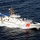 An aerial photograph of a U.S. Coast Guard vessel underway at high speed on open ocean waters. The white patrol boat bears the hull number 1114 and displays the distinctive USCG racing stripe (diagonal red and blue stripe with Coast Guard emblem) on its bow. The vessel is generating a significant white wake, indicating it is traveling at considerable speed.