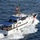 An aerial photograph of a U.S. Coast Guard vessel named 'Heriberto Hernandez' underway at speed on open blue ocean waters. The white cutter displays the distinctive USCG diagonal red stripe on its bow and is crewed by uniformed personnel visible on deck. The vessel appears to be a Fast Response Cutter (FRC) of the Sentinel-class.