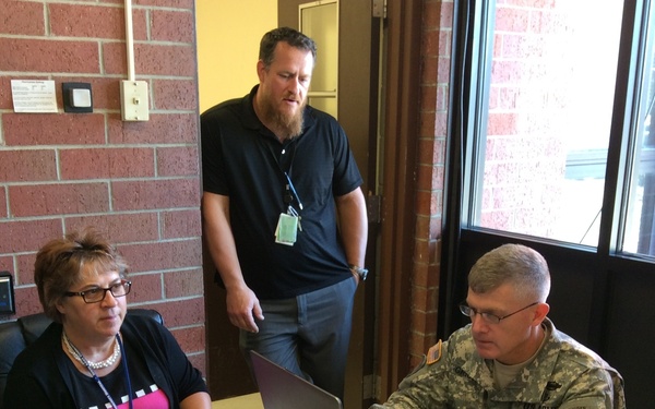 Three individuals are gathered around a desk in a brick-walled office or workspace. A U.S. Army soldier in ACU (Army Combat Uniform) is typing on a laptop, while a woman seated beside him observes, and a bearded man in civilian clothes stands behind them looking on. The setting appears to be a temporary operations or coordination center.