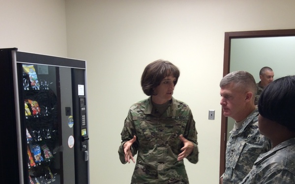 Three U.S. Army personnel in military uniforms are gathered in what appears to be a break room or common area near a snack vending machine. A female soldier in OCP (Operational Camouflage Pattern) uniform appears to be briefing or explaining something to two other soldiers wearing ACU (Army Combat Uniform) pattern. A fourth soldier is partially visible through a doorway in the background.