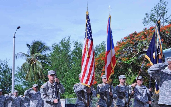 A U.S. Army color guard and formation of soldiers are conducting a military ceremony, likely a parade or formal review. Soldiers in ACU (Army Combat Uniform) digital camouflage are standing at salute while a color guard presents the U.S. flag, Puerto Rico flag, and at least one unit guidon. The setting appears to be a military installation in a tropical environment, consistent with Puerto Rico.
