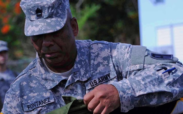 A U.S. Army soldier named Fontanez, wearing ACU (Army Combat Uniform) digital camouflage, is handling what appears to be a folded military guidon or ceremonial flag with gold fringe. The soldier displays rank insignia consistent with a senior NCO and is focused on the flag-handling task. A second soldier in ACU uniform is visible in the background, suggesting a military ceremony or formation.