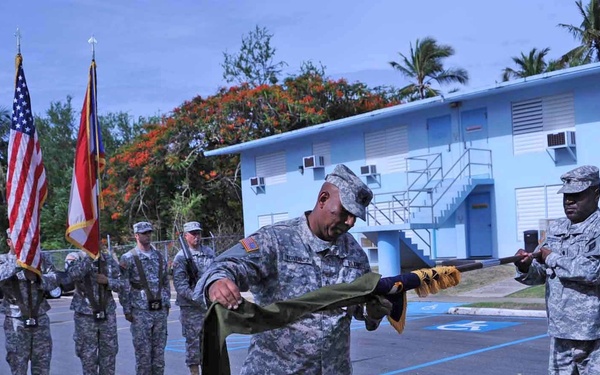 A U.S. Army military ceremony is taking place outdoors at what appears to be a tropical installation, likely in a Pacific or Caribbean location based on the palm trees and blue building architecture. A senior non-commissioned officer or officer is casing (wrapping) a unit guidon or colors in a green case, which is a formal military ritual typically associated with unit inactivation, deployment, or reactivation ceremonies. Soldiers in ACU (Army Combat Uniform) are standing in formation holding flags including the American flag.