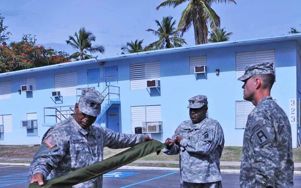 A group of U.S. Army soldiers in ACU (Army Combat Uniform) are conducting what appears to be a military ceremony in a parking lot. Two soldiers in the foreground are unfolding or presenting a green cloth/flag to a third soldier standing at attention. The setting appears to be a tropical military installation, indicated by palm trees and a light blue two-story building numbered '323' in the background.