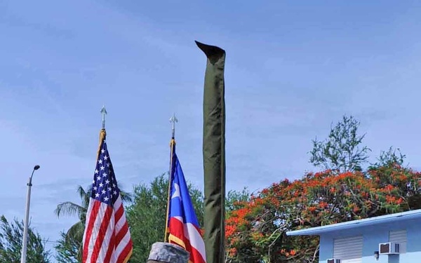 A military ceremony is taking place outdoors on a paved surface, likely a change of command or casing/uncasing of colors ceremony. Two U.S. Army soldiers in ACU (Army Combat Uniform) are at the center, exchanging or presenting a cased guidon/flag. The American flag and what appears to be the Puerto Rico flag are visible in the background, suggesting this may be a Puerto Rico National Guard unit.