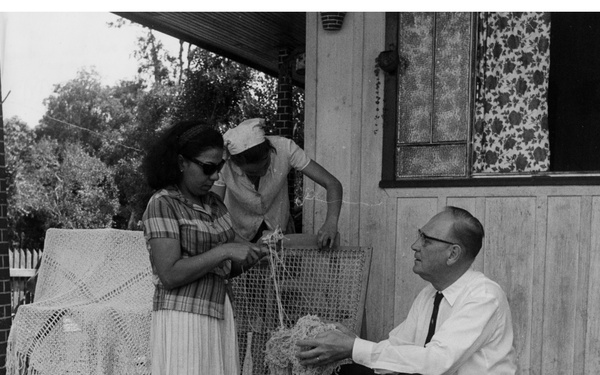 A black-and-white photograph showing three people on the front porch/steps of a wooden house, apparently examining or discussing a piece of handcrafted netting or lacework. Two women stand while a man sits on the steps holding what appears to be a loosely woven or frayed textile item. The setting appears to be a modest residential property, likely from the 1960s based on clothing styles and photographic quality.