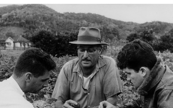 A black-and-white archival photograph showing three men crouching in an agricultural field, closely examining plant specimens or roots. The central figure wears a fedora hat and glasses, suggesting a possible expert or agronomist role. The setting appears to be a rural farming area with hills and a building visible in the background.