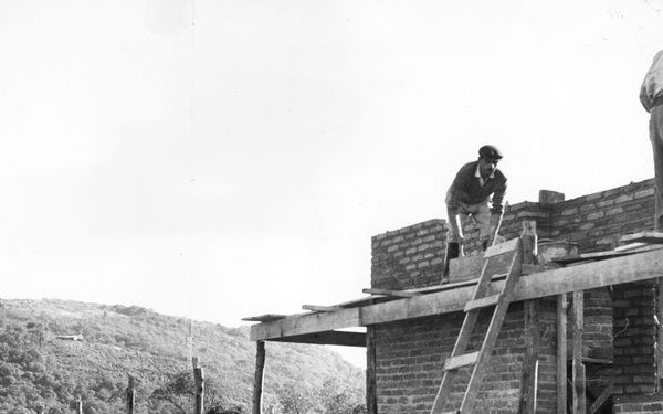 A black-and-white photograph depicting an active brick building construction site, likely from the mid-20th century. Multiple workers are engaged in masonry and structural work at various levels of the partially completed structure. The site is set against a hillside backdrop covered in dense vegetation.