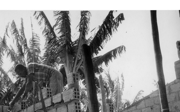 A black-and-white photograph showing a construction worker laying or working with concrete/cinder blocks on a partially built structure. The building is under active construction with walls reaching approximately two stories, surrounded by palm trees in the background. The photo appears to be taken from a low angle, looking up through wooden scaffolding or formwork.