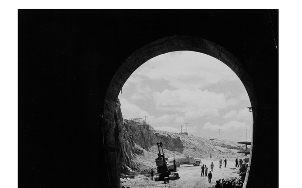 A black-and-white archival photograph taken from inside a tunnel looking outward through an arched opening toward an active construction or excavation site. The foreground shows water pooling on the tunnel floor with visible ripple rings, while silhouetted workers and heavy machinery are visible in the bright exterior. Rocky cut slopes and utility poles are visible in the background under a partly cloudy sky.