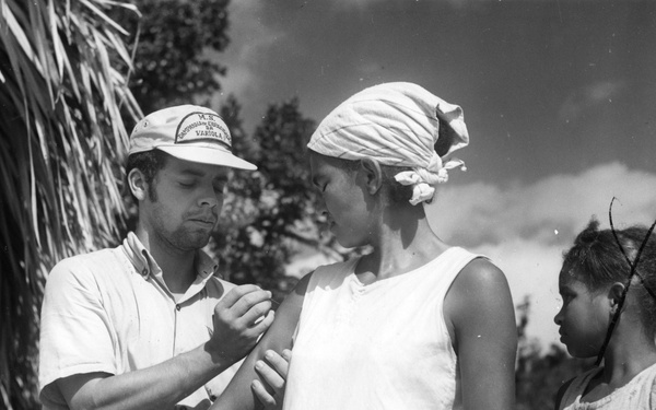 A black-and-white photograph showing a male health worker administering what appears to be a vaccination or medical examination to a young woman outdoors. The worker is wearing a cap with text that includes the word 'VARIOLA,' suggesting this is part of a smallpox eradication campaign. A second younger girl stands nearby observing the procedure.