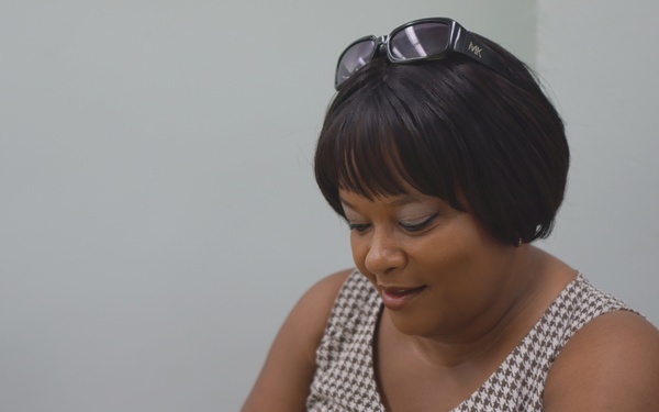 A woman is seated at a table engaged in a handcraft activity, likely crocheting or knitting with light blue and white yarn. She is wearing a houndstooth patterned sleeveless top with button details, sunglasses resting on her head, and a fitness tracker on her wrist. The setting appears to be an indoor room with a light-colored wall.