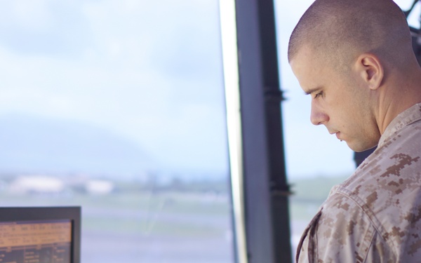 A military personnel member in desert camouflage uniform is working at what appears to be an air traffic control tower. The individual is focused on a control console equipped with multiple monitors and instrument panels, with a view of an airfield runway visible through large windows in the background. The setting strongly suggests a military airbase control tower environment.