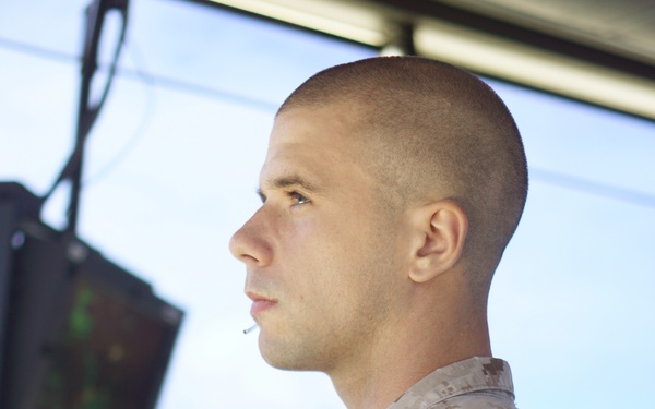 A young male military personnel wearing USMC MARPAT desert camouflage uniform is standing in what appears to be an air traffic control tower. He is facing to the left in profile view, with a pen or stylus held in his mouth. Multiple radar/computer screens are visible in the background.