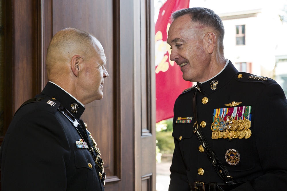 Two senior U.S. Marine Corps officers in dress blue uniforms are engaged in a friendly conversation near a doorway. Both men display extensive medal and ribbon arrays indicating high-ranking positions and long service careers. A red Marine Corps flag with gold eagle, globe, and anchor emblem is visible in the background.