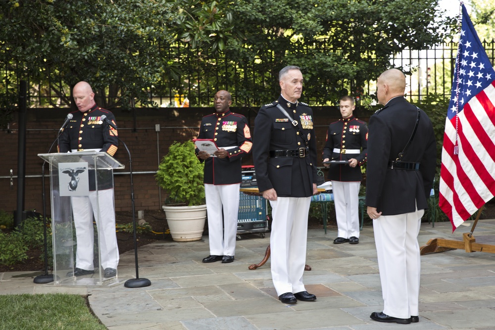 An outdoor military ceremony featuring United States Marine Corps personnel in dress uniforms. The event appears to be a formal occasion with a podium bearing the Marine Corps Eagle, Globe, and Anchor emblem, an American flag, and multiple Marines of varying ranks assembled on a stone patio. The setting includes a garden or courtyard with trees, an iron fence, and audio equipment in the background.