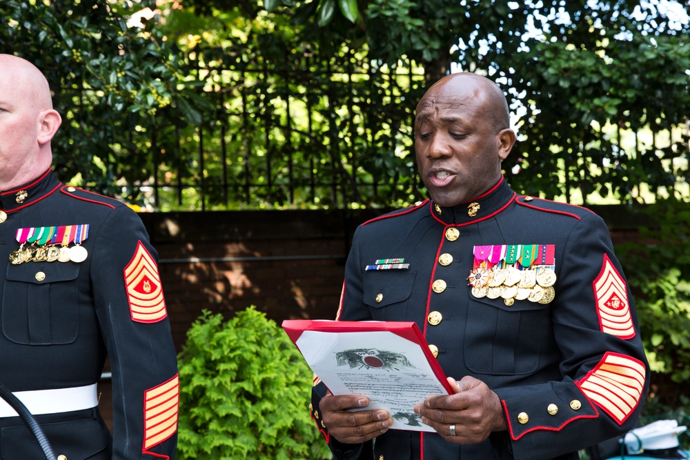 Two United States Marine Corps personnel in dress blue uniforms are participating in what appears to be a formal military ceremony outdoors. The central figure, a senior non-commissioned officer, is reading from a document held in a red-bordered presentation folder. The setting includes a wrought iron fence and lush green foliage in the background.