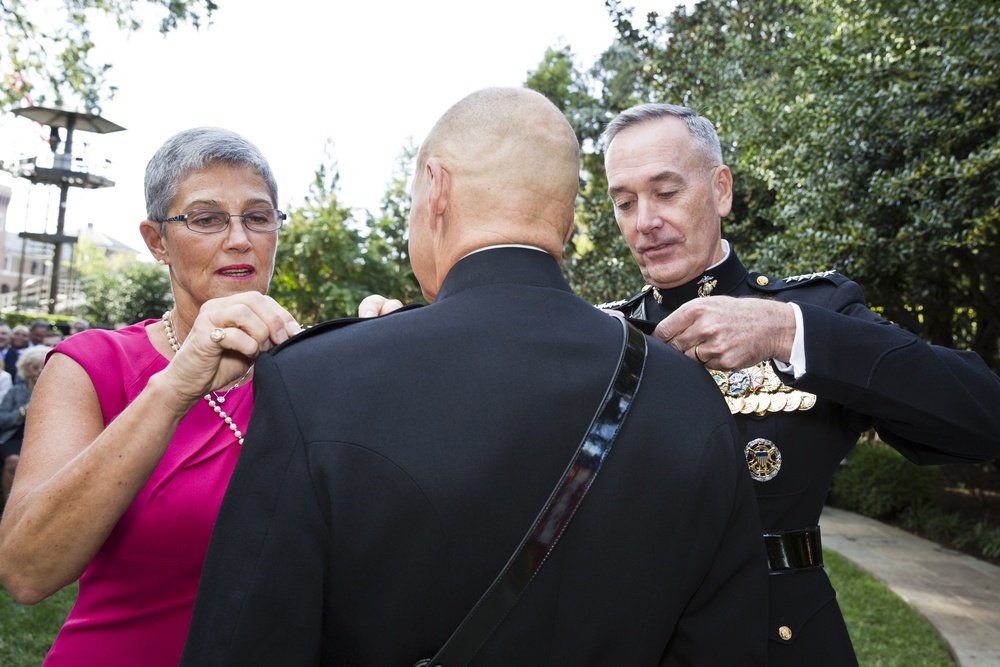 A military promotion or decoration ceremony taking place outdoors, where a senior military officer in dress uniform and a woman in a pink dress are pinning insignia or rank devices onto the shoulders of a third individual whose back is to the camera. The setting appears to be a formal garden or courtyard with trees and other attendees visible in the background. This is consistent with a traditional military pinning ceremony.