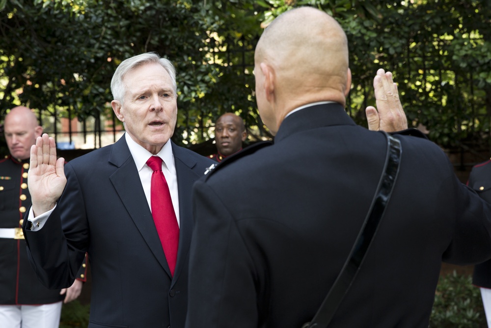 A formal oath-taking ceremony is taking place outdoors, with a white-haired man in a dark suit and red tie raising his right hand while facing another individual who is also raising their right hand. Several individuals in dark military dress uniforms are visible in the background. The setting appears to be an outdoor courtyard or garden area with iron fencing and green foliage.