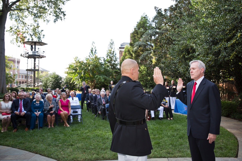 An outdoor swearing-in or oath-taking ceremony is taking place on a grass lawn, with a uniformed military officer (facing away from camera) administering an oath to a civilian man in a dark suit with a red tie. Both individuals have their right hands raised. A seated audience of approximately 30-40 attendees observes the ceremony, with additional uniformed military personnel visible in the background.