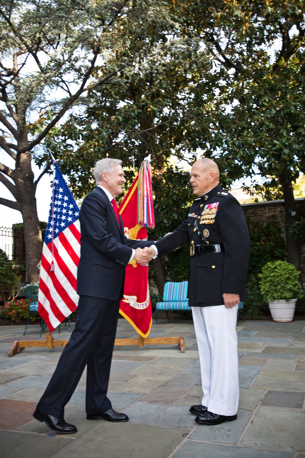 Two men are shaking hands outdoors on a stone patio, with an American flag and what appears to be a United States Marine Corps flag visible in the background. One man is wearing a dark civilian suit with a red tie, while the other is dressed in a formal U.S. Marine Corps dress blue uniform with multiple medals and ribbons. The setting appears to be a formal ceremonial or official occasion.