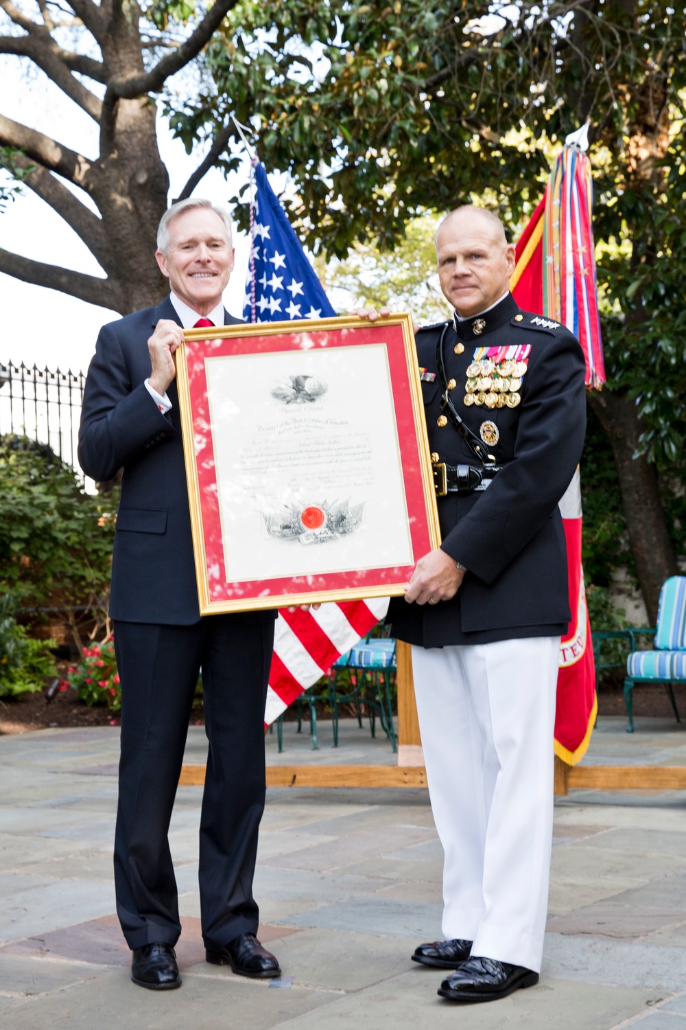 Two men stand outdoors on a stone patio holding a framed official document with red matting and a gold frame. One man is in civilian attire (dark suit, red tie) and the other is in a U.S. Marine Corps dress blue uniform with numerous medals and white trousers. The setting appears to be a formal military ceremony with American and military flags visible in the background.