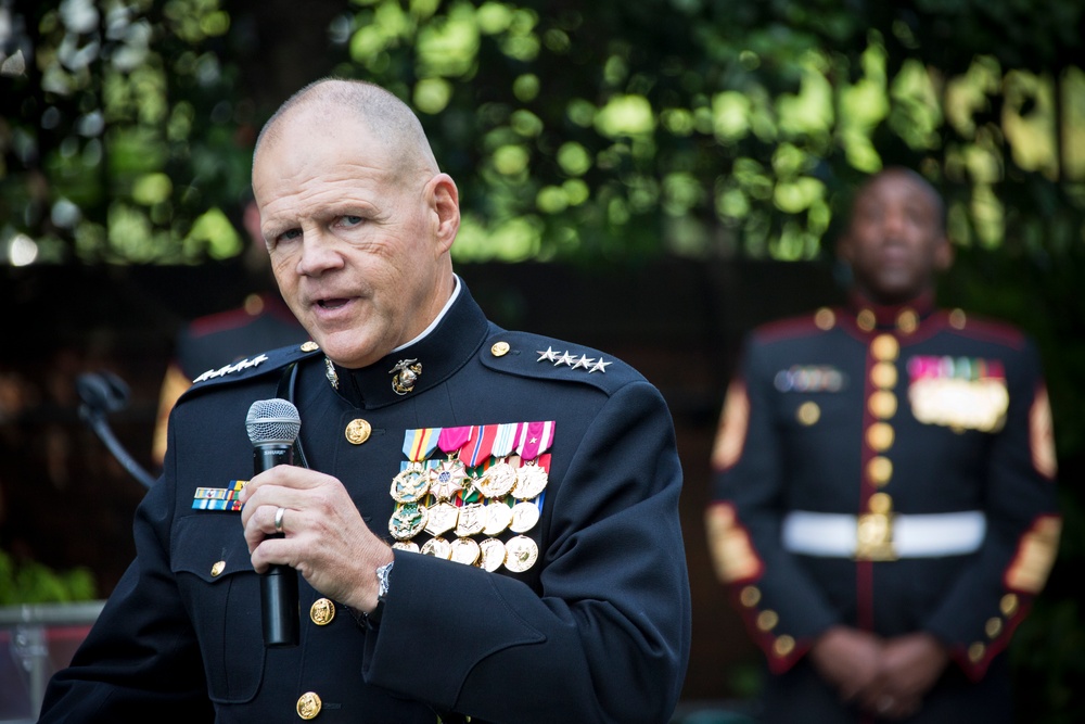 A senior U.S. Marine Corps officer in dress blues is speaking into a microphone at what appears to be an outdoor formal event. He displays four stars on his shoulder boards indicating the rank of General, and wears an extensive array of medals and decorations on his chest. A second Marine in dress uniform stands in the background.