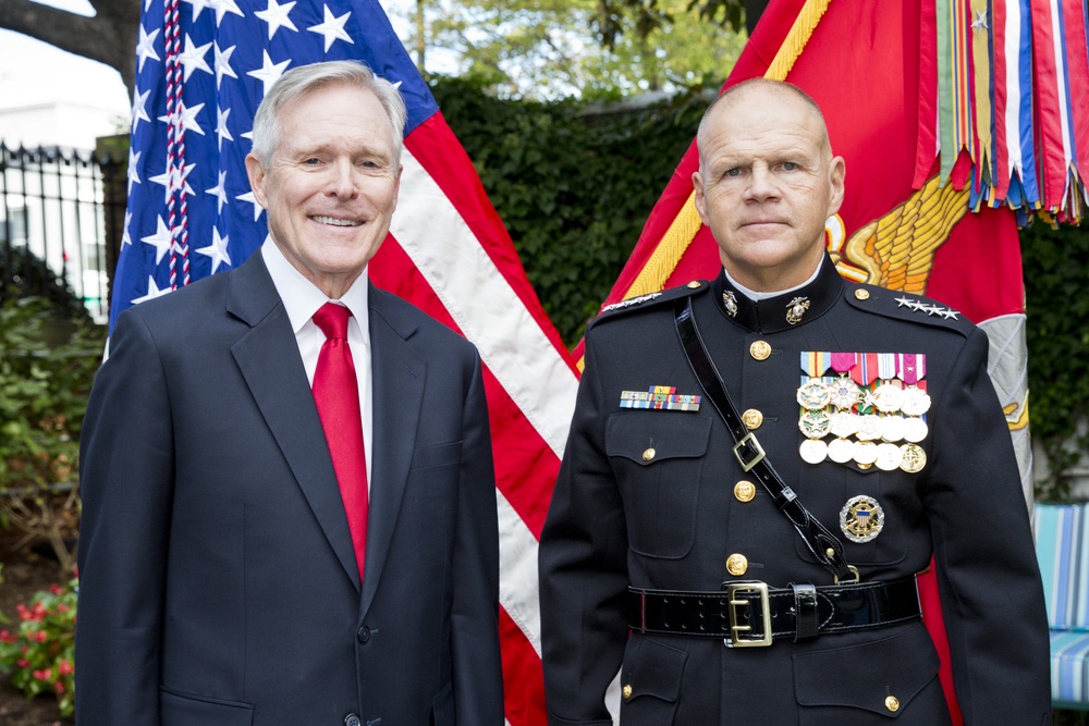 Two men pose formally in front of an American flag and what appears to be a Marine Corps flag. The man on the left wears a dark civilian suit with a red tie, while the man on the right wears a U.S. Marine Corps dress uniform adorned with numerous medals, ribbons, and rank insignia indicating a high-ranking officer. The setting appears to be an outdoor official or ceremonial event.