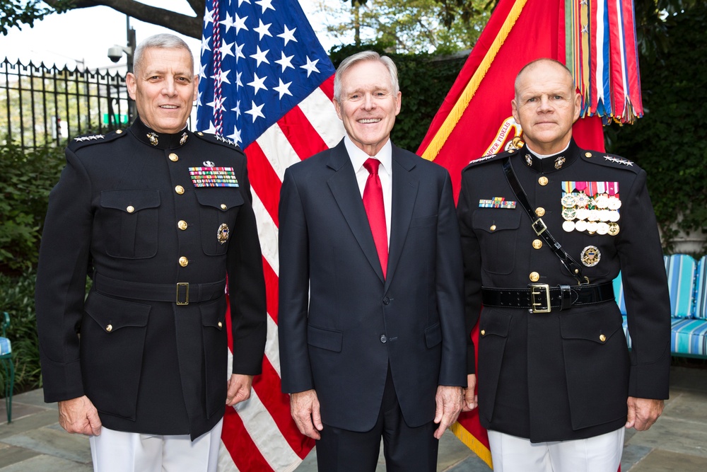 Three men pose formally in front of an American flag and a red military ceremonial flag. Two of the men are wearing U.S. Marine Corps dress blue uniforms with medals and rank insignia, while the man in the center is wearing a civilian dark suit with a red tie. The setting appears to be an outdoor ceremonial or official event.