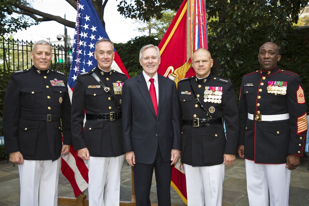 Five individuals pose formally for a group photograph outdoors, with four wearing U.S. Marine Corps dress uniforms adorned with medals and insignia, and one civilian in a dark suit with a red tie. An American flag and a red U.S. Marine Corps flag are visible in the background. The setting appears to be an official military or government ceremony.