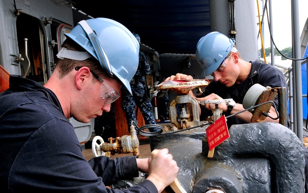 Two workers wearing blue hard hats and safety glasses are performing maintenance work on large industrial pipes and valves on what appears to be a naval vessel. A third person in Navy digital camouflage uniform is visible in the background. The workers are using hand tools, including what appears to be a wooden mallet, to work on corroded pipe fittings and valve assemblies.