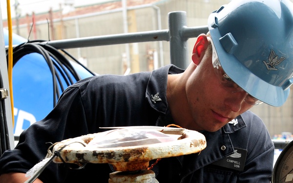 A U.S. Navy sailor identified by name tape as 'CAMPBELL, USN' is performing maintenance work on a heavily corroded valve or pipe fitting. He is wearing a light blue hard hat with a petty officer insignia and dark navy working uniform. The work appears to be taking place aboard a naval vessel or at a naval facility.