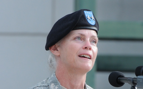 A U.S. Army Major General is speaking at a podium with microphones during what appears to be an official military ceremony or event. The officer is wearing Army Combat Uniform (ACU) with a black beret bearing a unit flash, and displays two-star rank insignia on the collar. The name tape on the uniform reads 'LIN' and the branch tape reads 'U.S. ARMY'.