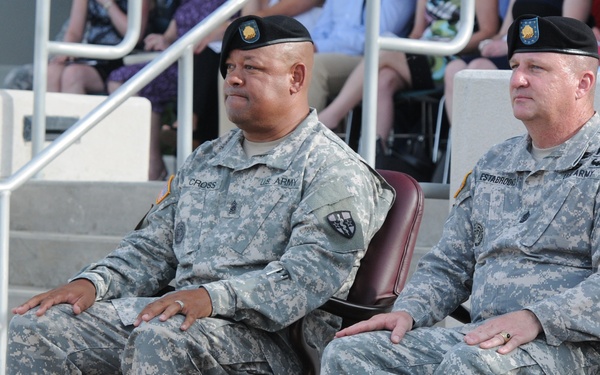 Two U.S. Army soldiers in ACU (Army Combat Uniform) are seated in formal chairs, likely during an official military ceremony or event. Both wear black berets with insignia and have name tapes visible on their uniforms. An audience is visible seated on steps in the background.