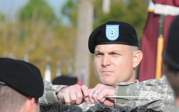 A U.S. Army soldier wearing a black beret with a blue flash insignia and ACU (Army Combat Uniform) is conducting what appears to be a military ceremony or formation. The name tape on his uniform reads 'GREEN' and 'U.S. ARMY'. He is holding what appears to be a baton or pointer while addressing soldiers in the foreground.