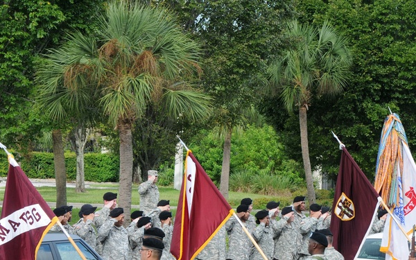 A formal U.S. Army military ceremony is taking place outdoors, likely on a military installation. Soldiers in ACU (Army Combat Uniform) are rendering salutes while color guard members hold multiple unit guidons and the U.S. national flag. The ceremony appears to be a passing of honors, possibly a change of command or dignitary arrival, with vehicles passing through a formation.