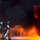 A team of firefighters wearing full silver aluminized proximity suits and breathing apparatus are actively combating a large, intense fire. The lead firefighter is directing a water or foam stream toward the flames while teammates provide support from behind. Several additional personnel are visible in the background observing or standing by.