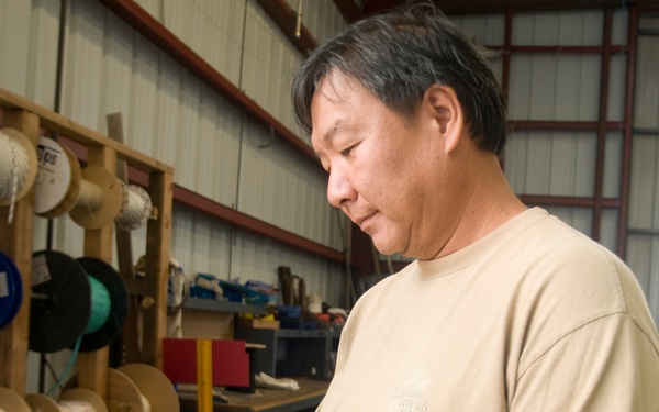A middle-aged man is standing in what appears to be an industrial workshop or supply warehouse, holding and reviewing a tablet device. The background shows organized shelving units stocked with various spools of wire, rope, and cable in multiple colors. The setting suggests a trade or electrical supply environment where the tablet may be used for inventory or work orders.
