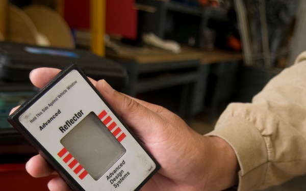 Two hands are holding a pair of devices manufactured by Advanced Design Systems: an 'Advanced Reflector' unit and an 'Advanced Light Transmittance Meter'. The meter's LCD display shows a reading of '100.0' percent transmittance, indicating the devices are being demonstrated or calibrated without a window between them. The setting appears to be an automotive workshop or garage environment.