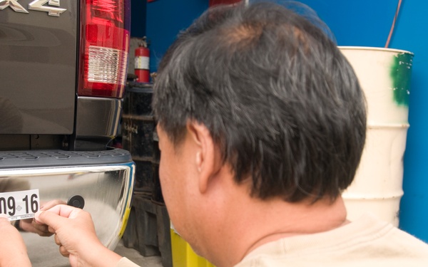 A man is affixing a vehicle permit sticker to the rear bumper of a truck, likely in a garage or service area. The blue sticker reads 'VEHICLE PERMIT - CITY &amp; COUNTY OF HONOLULU' with an expiration month of July and a partial number visible as '09 16'. The setting appears to be an automotive service or registration facility.