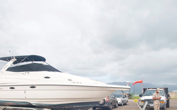 A large white Regal motorboat is loaded on a multi-axle flatbed trailer in a paved lot, likely near a boat launch facility. Two individuals are present — one in military-style clothing standing near the rear of the truck, and another crouching at the rear of the trailer. Mountains and overcast skies are visible in the background.