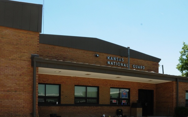 The image shows the exterior of a Kansas National Guard building, a brick structure with a covered entrance and signage above the door. Two individuals in military-style uniforms are visible near the entrance, with one actively sweeping or working on a concrete surface. The area in front of the building shows signs of recent landscaping or construction work, with disturbed soil visible.
