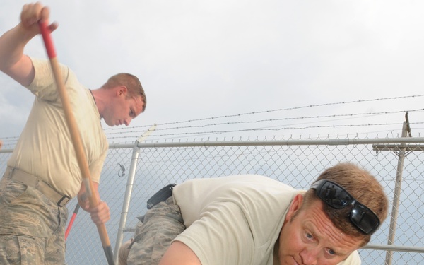 Multiple individuals wearing military camouflage uniforms are engaged in concrete or mortar work near a chain-link fence topped with barbed wire. The foreground subject, wearing a tan t-shirt and sunglasses on his forehead, is actively spreading or smoothing wet concrete/mortar with his hands and a tool. Additional personnel in the background are using shovels and hand tools to assist with the construction task.