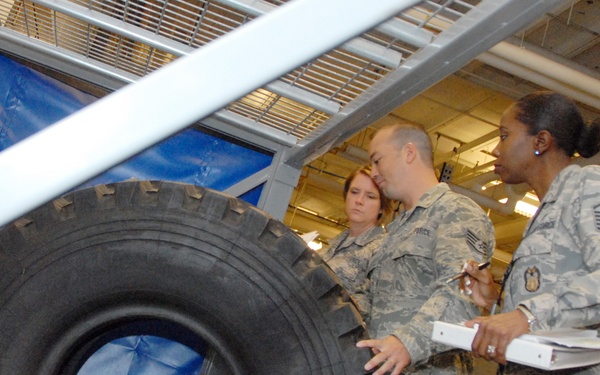 Three individuals in US Air Force camouflage uniforms (ABU pattern) are gathered in what appears to be a military maintenance hangar or facility. They appear to be conducting an inspection or review, with at least one person holding a binder/clipboard and another holding a pen. A large vehicle tire is prominently visible in the foreground.
