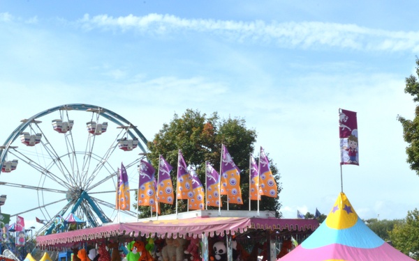 A busy outdoor carnival or state fair scene photographed on a clear, sunny day. Multiple game booths adorned with stuffed animal prizes, colorful tents, and decorative flags are visible, with a large Ferris wheel prominent in the background. Numerous fairgoers of various ages are walking through the midway area.
