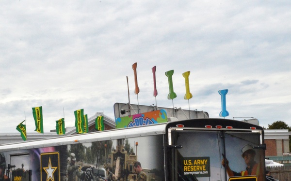 A U.S. Army Reserve recruitment trailer is parked at what appears to be a fairground or outdoor event. The trailer features full wrap graphics depicting soldiers in combat gear, Army logos, and recruitment messaging. The surrounding area shows carnival/fair attractions, colorful flags, and vendor tents.