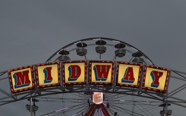 A carnival or state fair midway photographed at dusk, featuring a large illuminated 'MIDWAY' sign mounted on an archway in front of a Ferris wheel. The scene is bustling with fairgoers and lit by colorful lights against an overcast evening sky. Multiple carnival attractions, vendor tents, and a ticket booth are visible in the background.