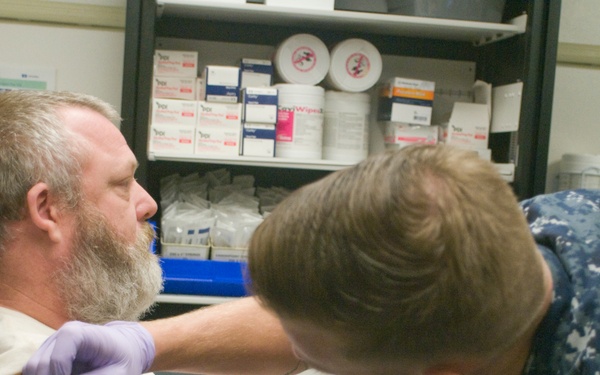 A medical professional wearing U.S. Navy digital camouflage uniform and purple latex gloves is administering an injection or performing a medical procedure on a bearded middle-aged man's upper arm. The patient is seated in a blue medical chair with his sleeve rolled up, appearing calm. The setting is a clinical medical room with a supply shelf stocked with medical materials in the background.