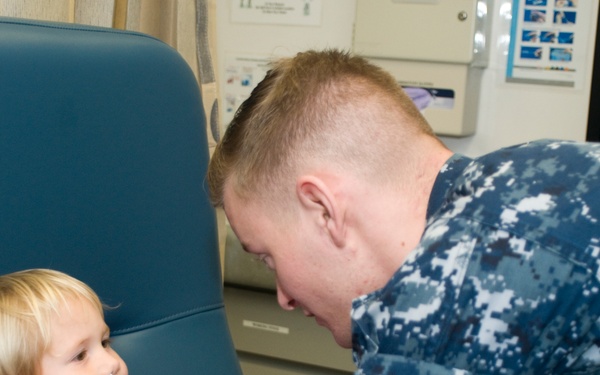 A person wearing US Navy digital camouflage (NWU) uniform and purple latex gloves is administering what appears to be a nasal spray or swab to a young blonde child seated in a medical/examination chair. The setting appears to be a military medical clinic or sick bay. The child is wearing a red shirt and appears calm during the procedure.