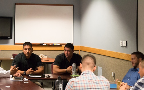 A group of approximately six to seven men are seated around a conference table in a meeting room, engaged in discussion. The man in a white long-sleeve shirt on the left appears to be speaking or leading the conversation. The setting suggests a professional or educational meeting environment.